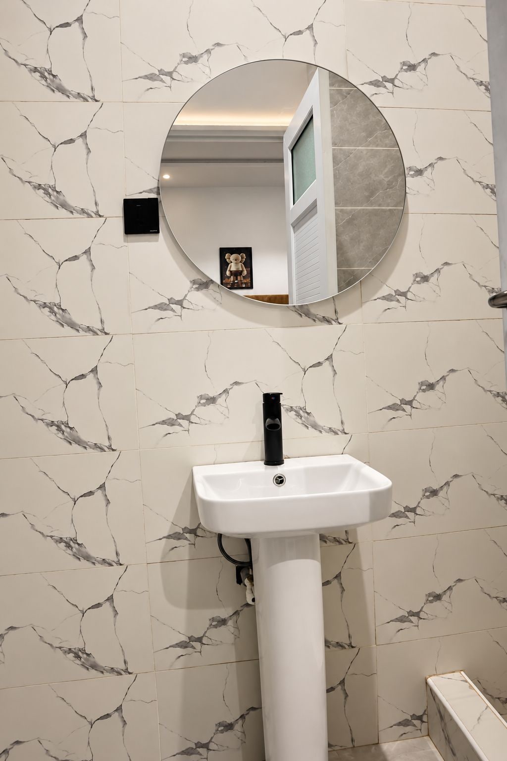 Casa de banho com parede de mármore branco, espelho redondo e lavatório pedestal branco.