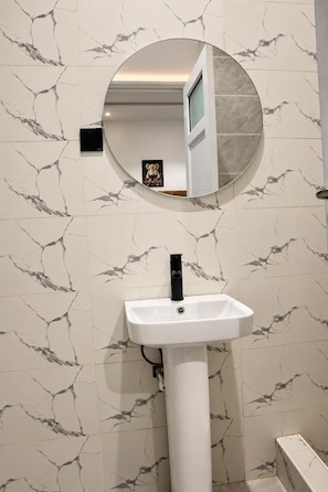 Casa de banho com parede de mármore branco, espelho redondo e lavatório pedestal branco.