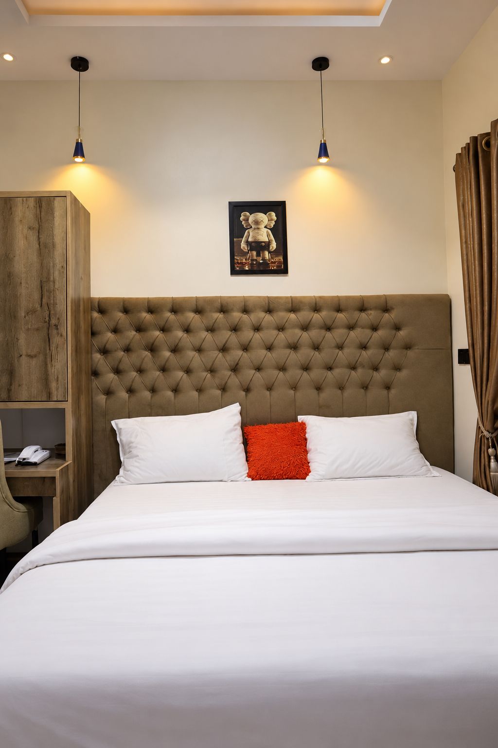 Quarto com cabeceira estofada, cama com almofadas brancas e vermelhas, guarda-roupa de madeira e quadro emoldurado na parede.