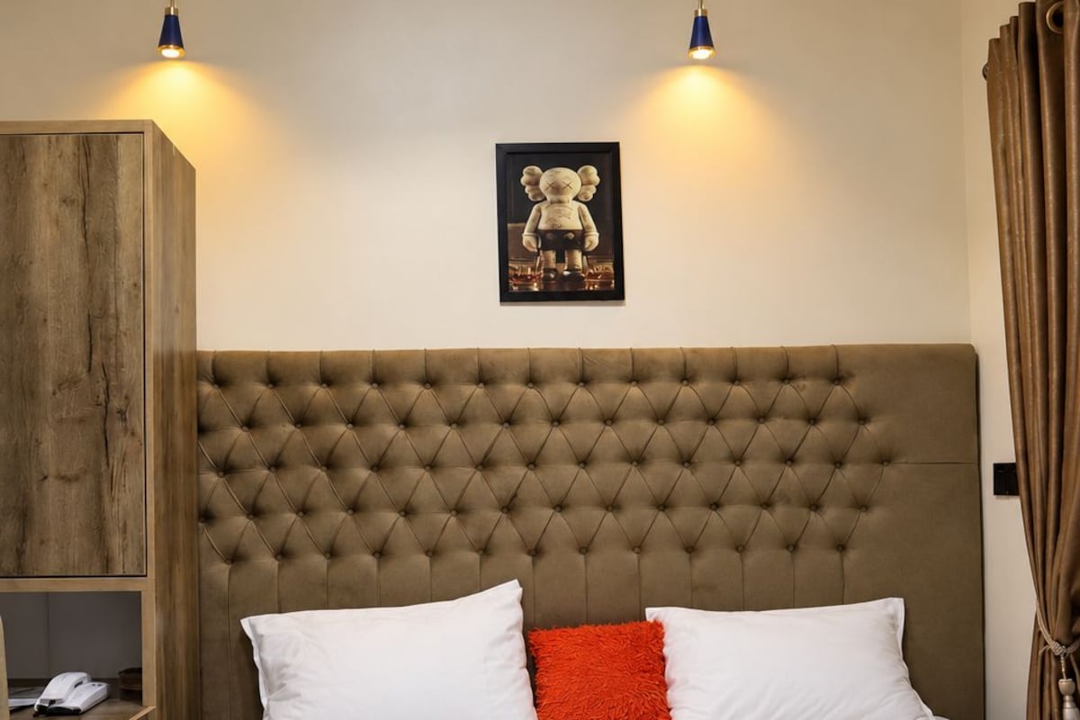 Quarto com cabeceira estofada, cama com almofadas brancas e vermelhas, guarda-roupa de madeira e quadro emoldurado na parede.