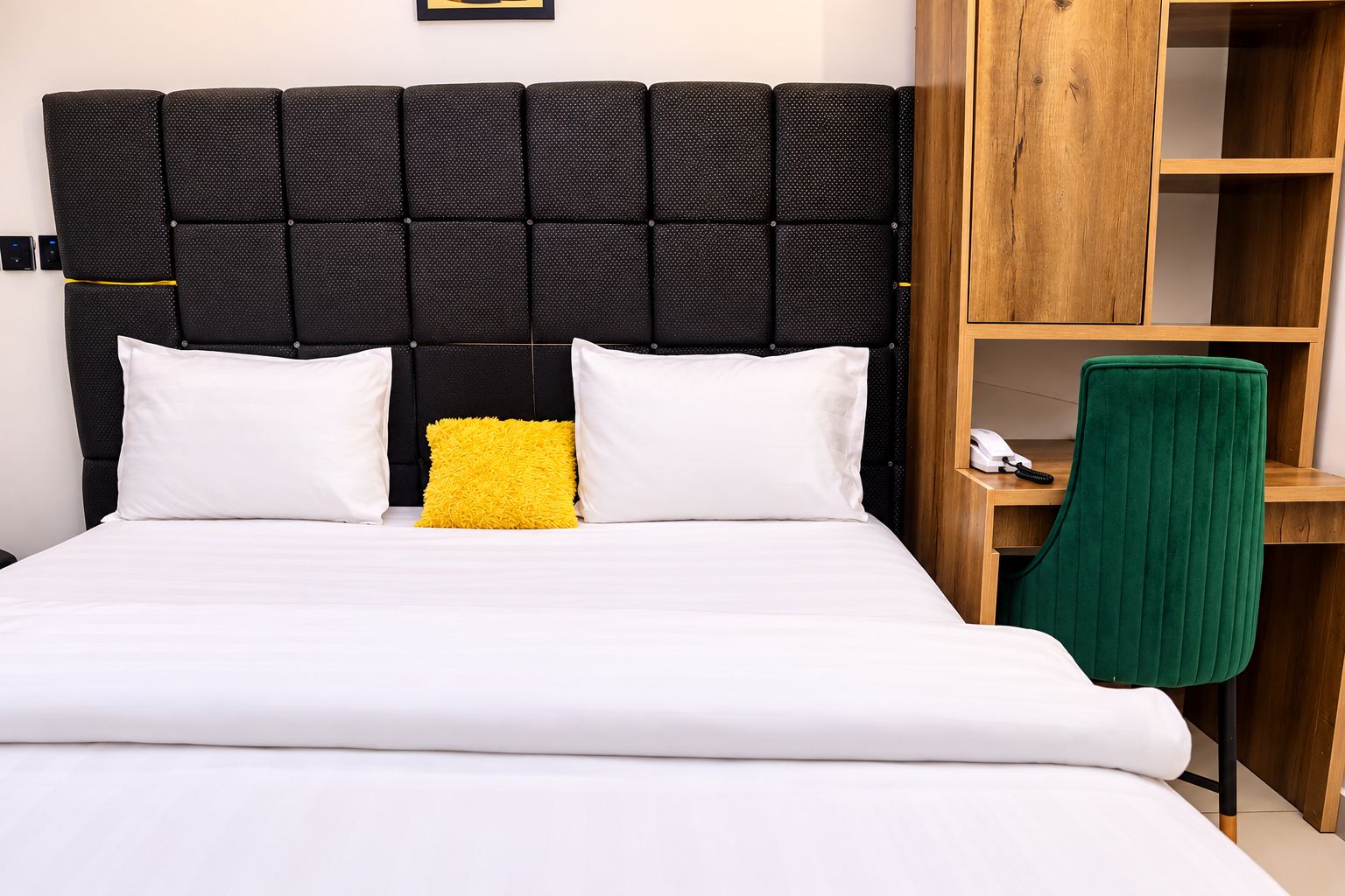 Cama com cabeceira preta, roupa de cama branca e almofada amarela de destaque.