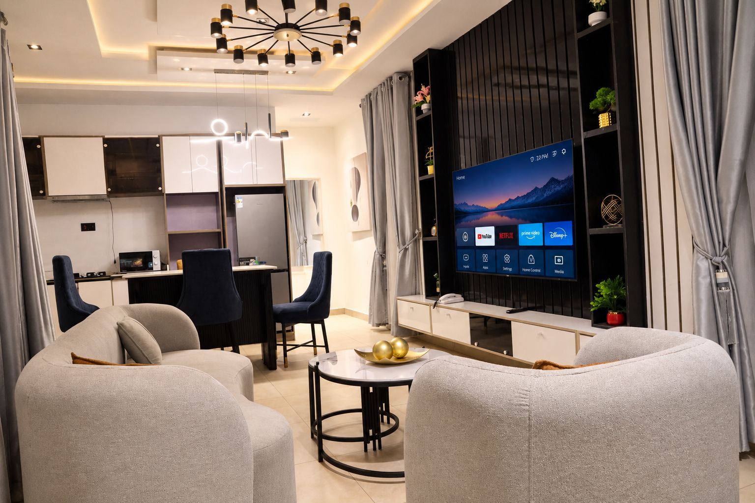 A modern living room with a flat-screen TV, seating area, and a kitchenette.