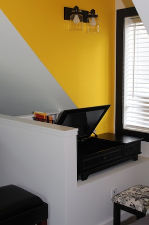 A room with a sloped ceiling, a black desk with a computer, a small bench, and a window with blinds.