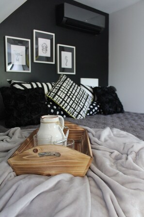A neatly made bed with a wooden tray containing a pitcher, glasses, and a spoon, set on a grey blanket.