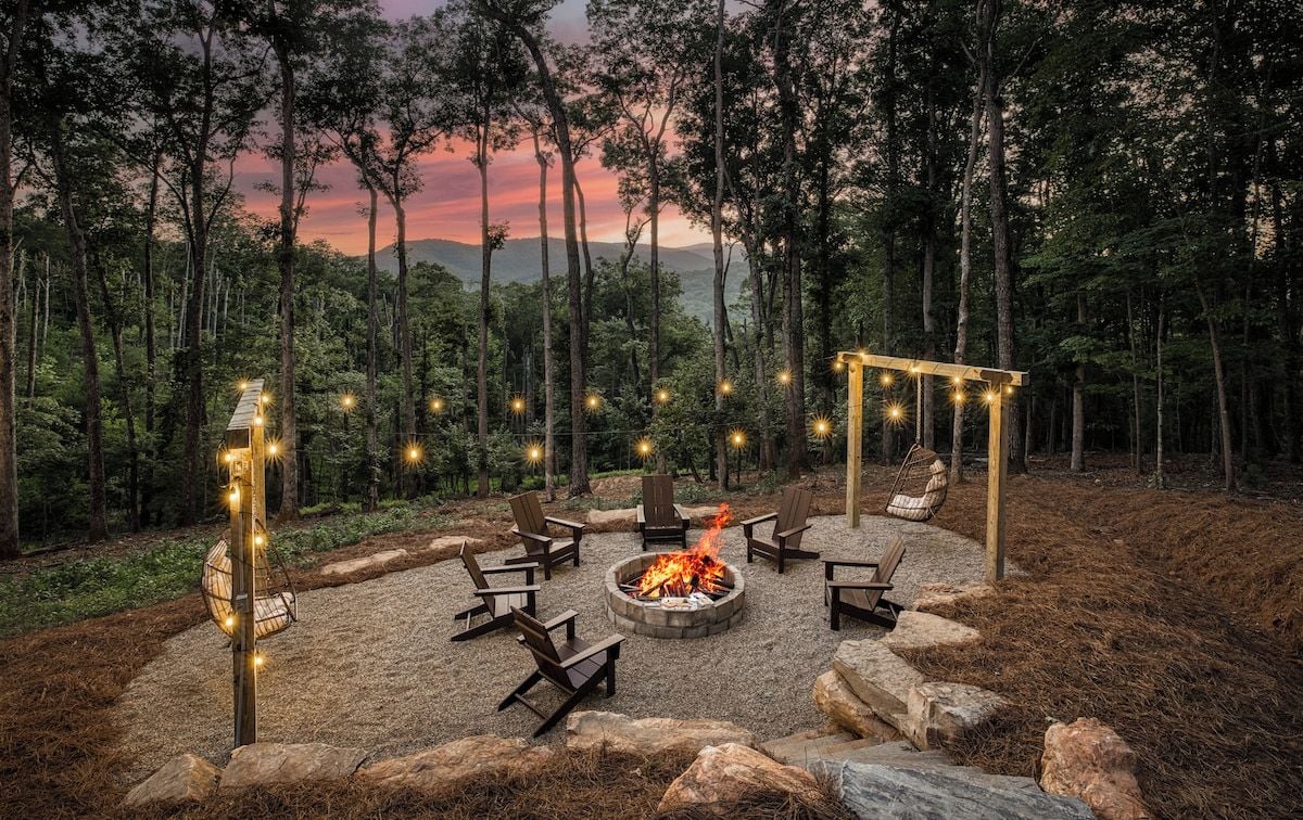 An outdoor seating area with a fire pit surrounded by chairs, set in a forested landscape.