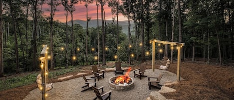 An outdoor seating area with a fire pit surrounded by chairs, set in a forested landscape.