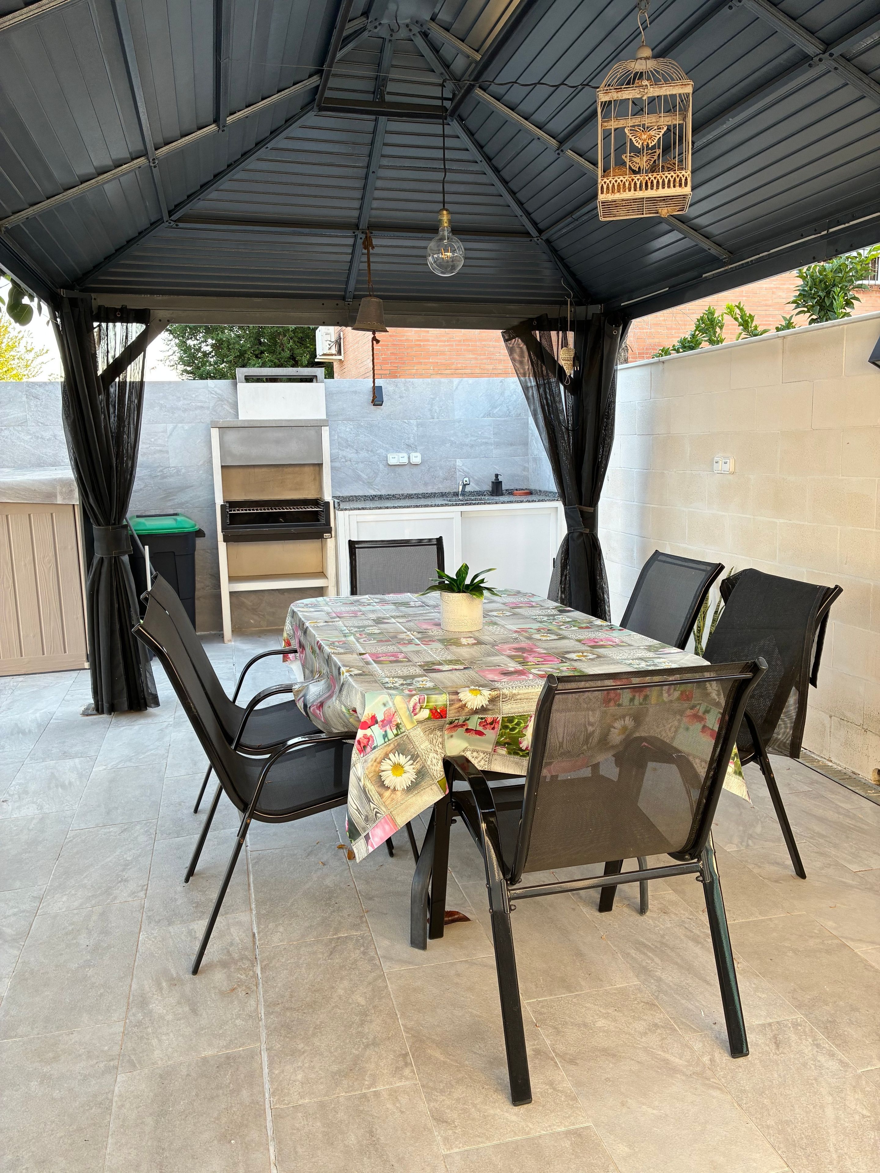 Eine Terrasse im Freien mit einem Esstisch, umgeben von Stühlen, einer Hängelampe und einem dekorativen Vogelkäfig.