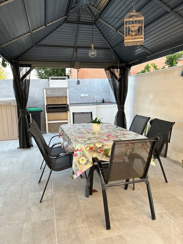 An outdoor patio with a table set for dining, surrounded by chairs, a hanging light fixture, and a decorative birdcage.