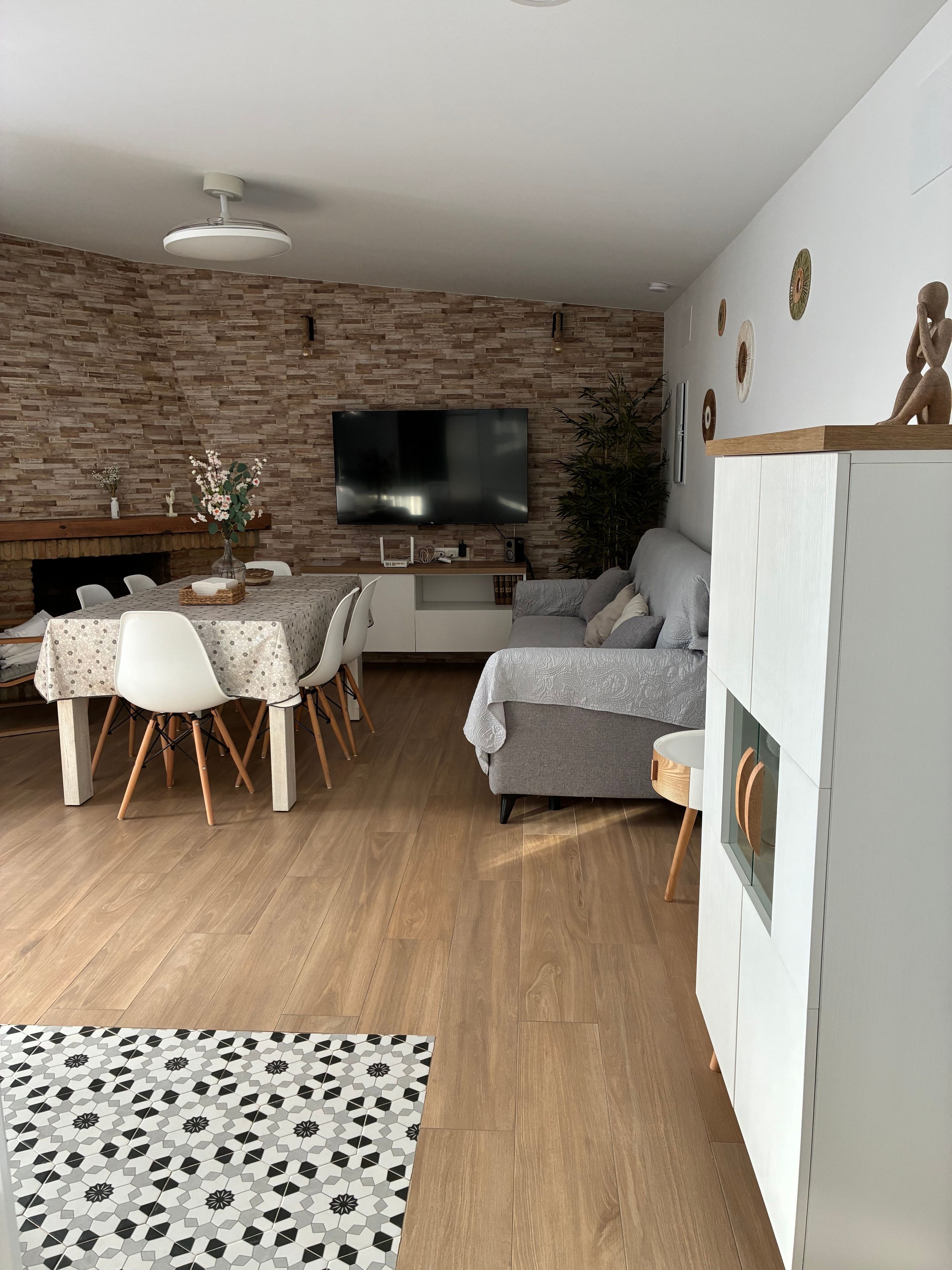 Ein modernes Wohnzimmer mit Backsteinwand, Flachbildfernseher, grauem Sofa, Esstisch mit Stühlen und einem weißen Schrank.