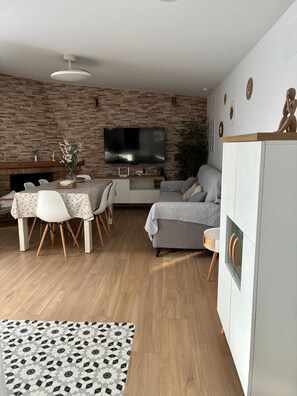 A modern living room with a brick wall, a flat-screen TV, a grey sofa, a dining table with chairs, and a white cabinet.