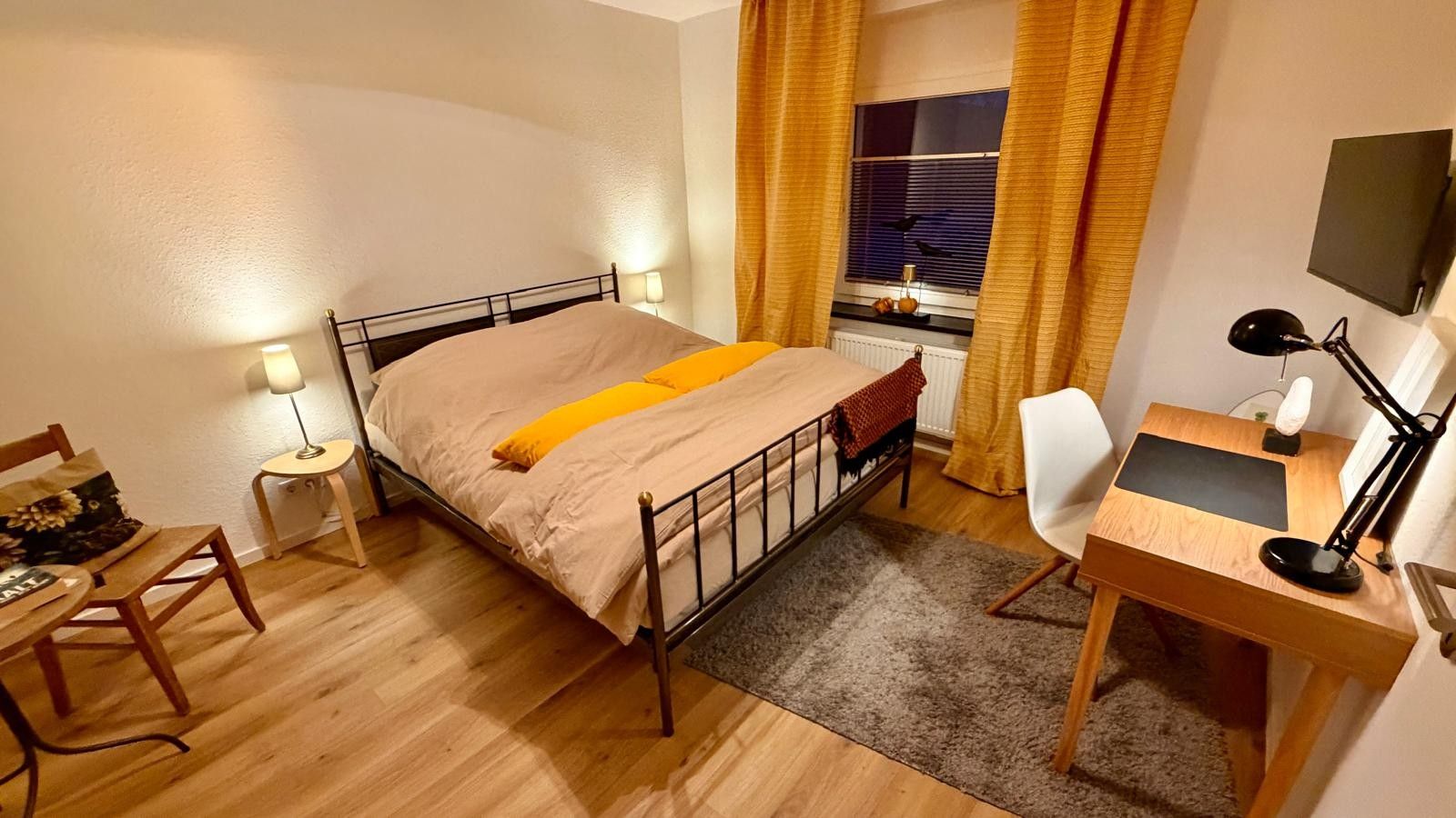 A bedroom with a bed, bedside table, chair, desk, and lamp.