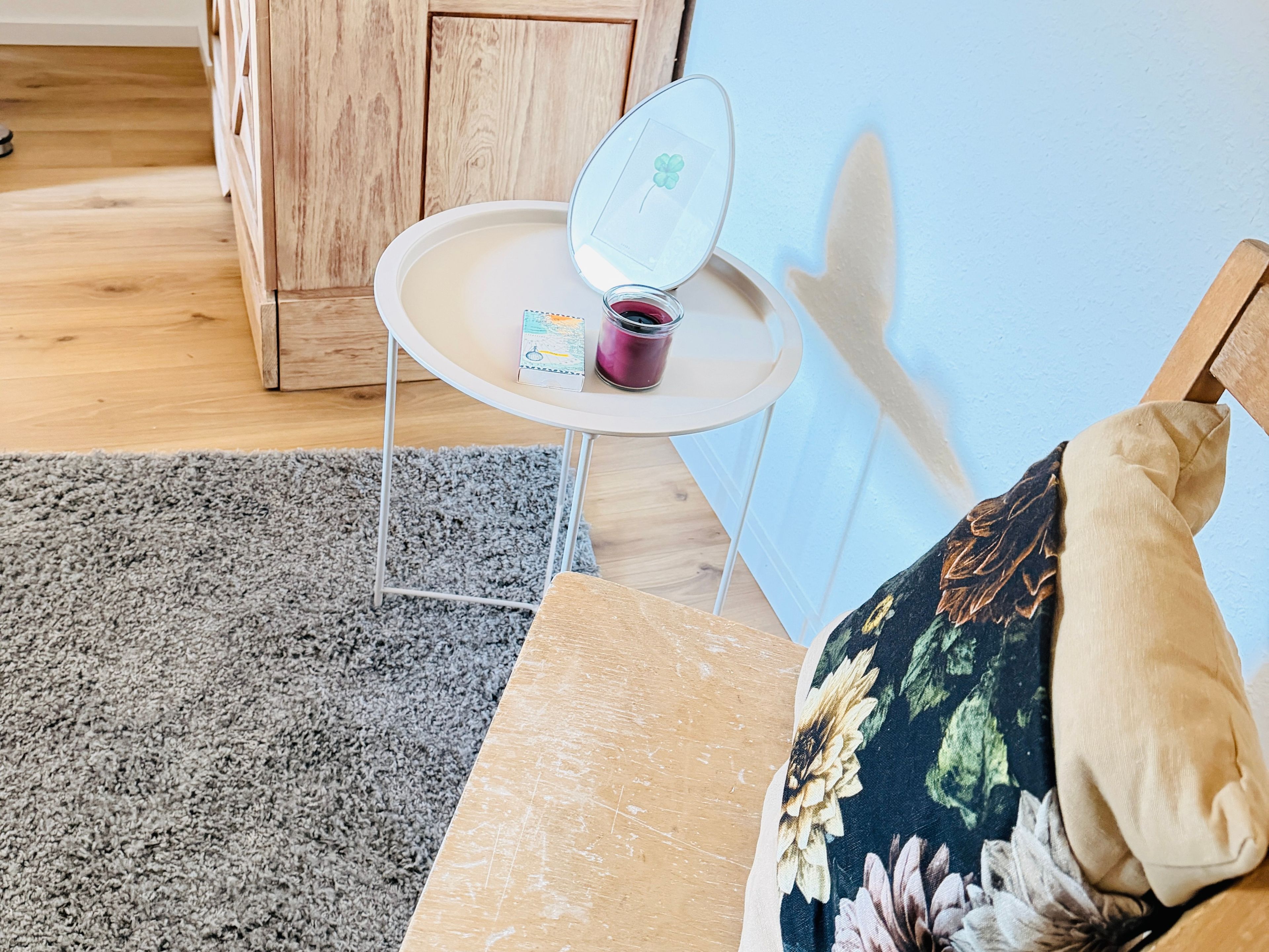 Ein Zimmer mit einem Holzkabinett, einem kleinen runden Tisch mit einer Kerze, einem grauen Teppich und einem blumigen Kissen auf einem Stuhl.