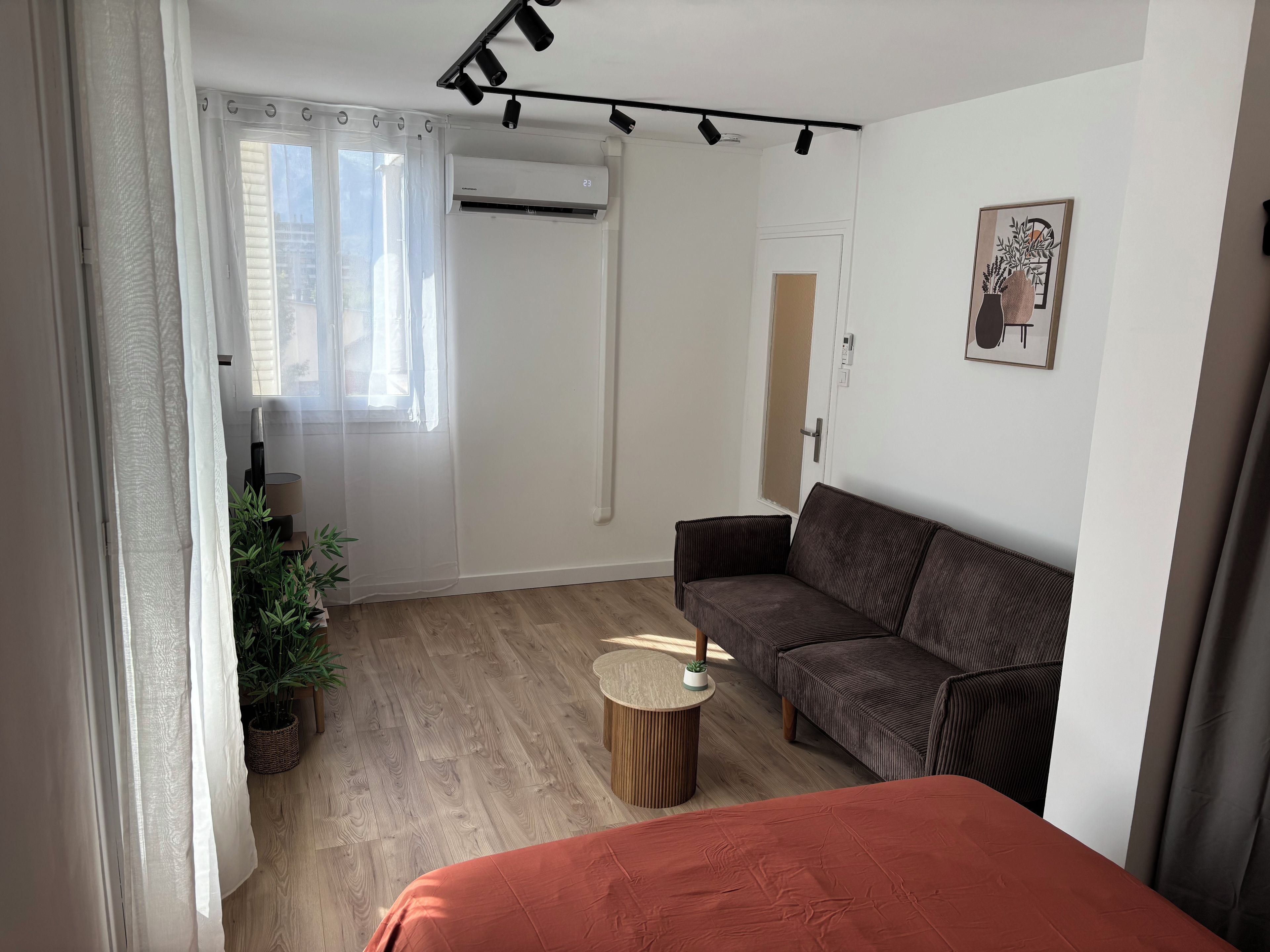 Una habitación con un sofá marrón, una mesa redonda de madera, una planta en maceta y una ventana con cortinas traslúcidas.