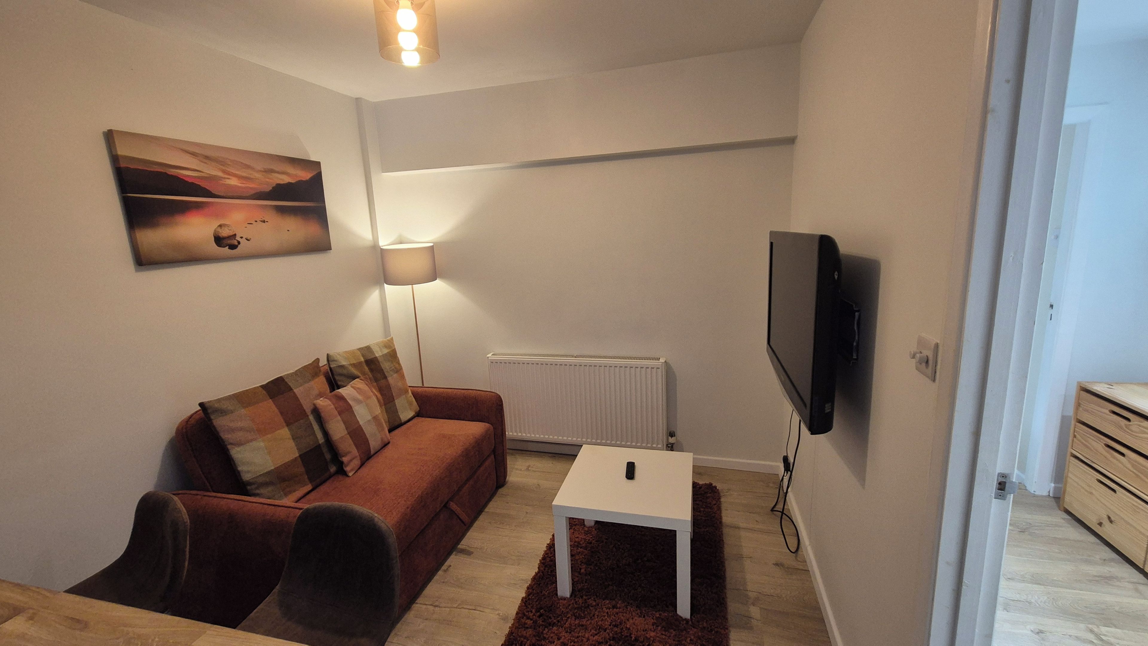 Ein Wohnzimmer mit einer Couch, einem Tisch, einer Lampe, einem Fernseher und einem Bild an der Wand.