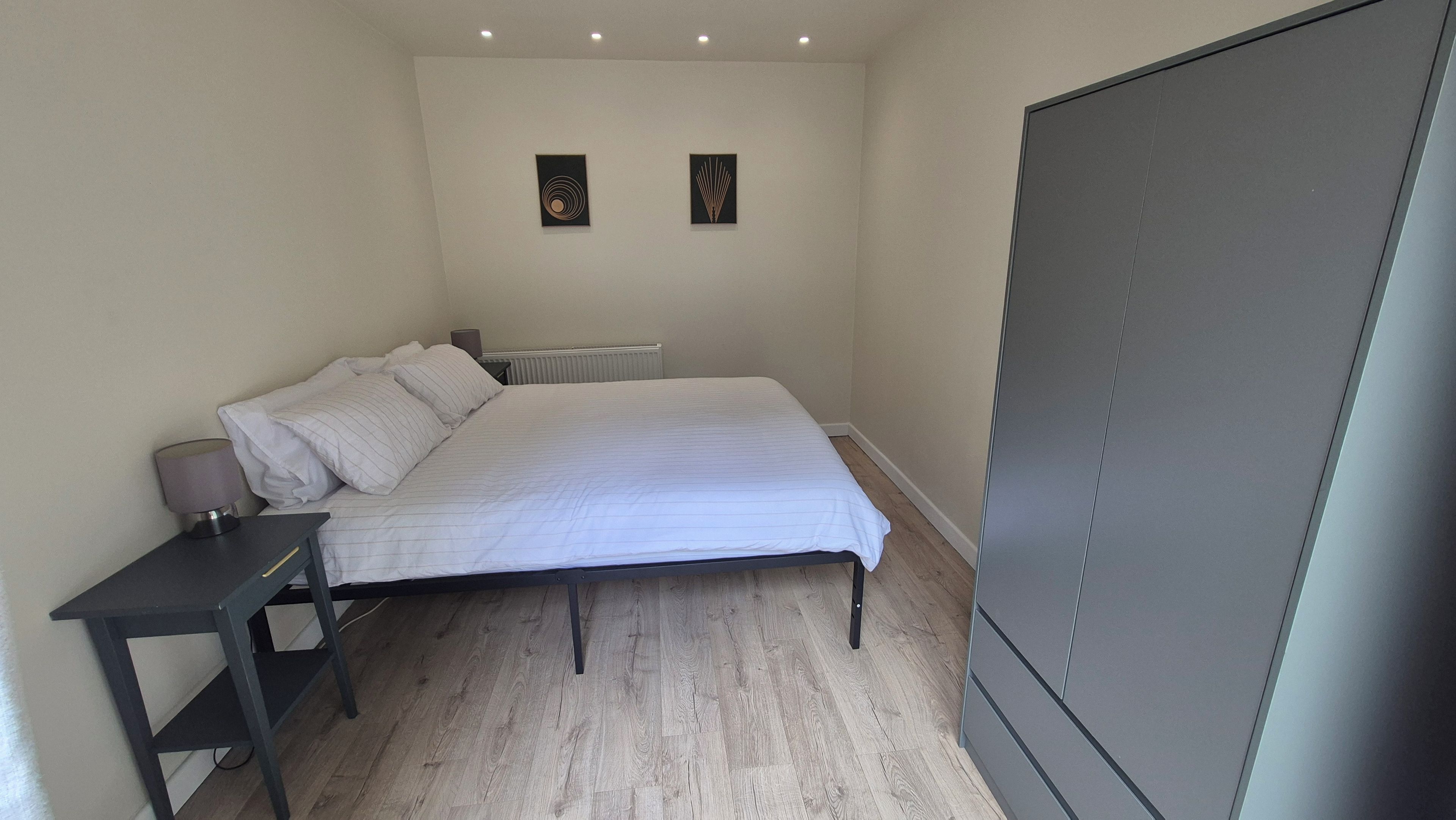 Een slaapkamer met een bed, nachtkastje, lamp, radiator en twee aan de muur hangende kunstwerken.