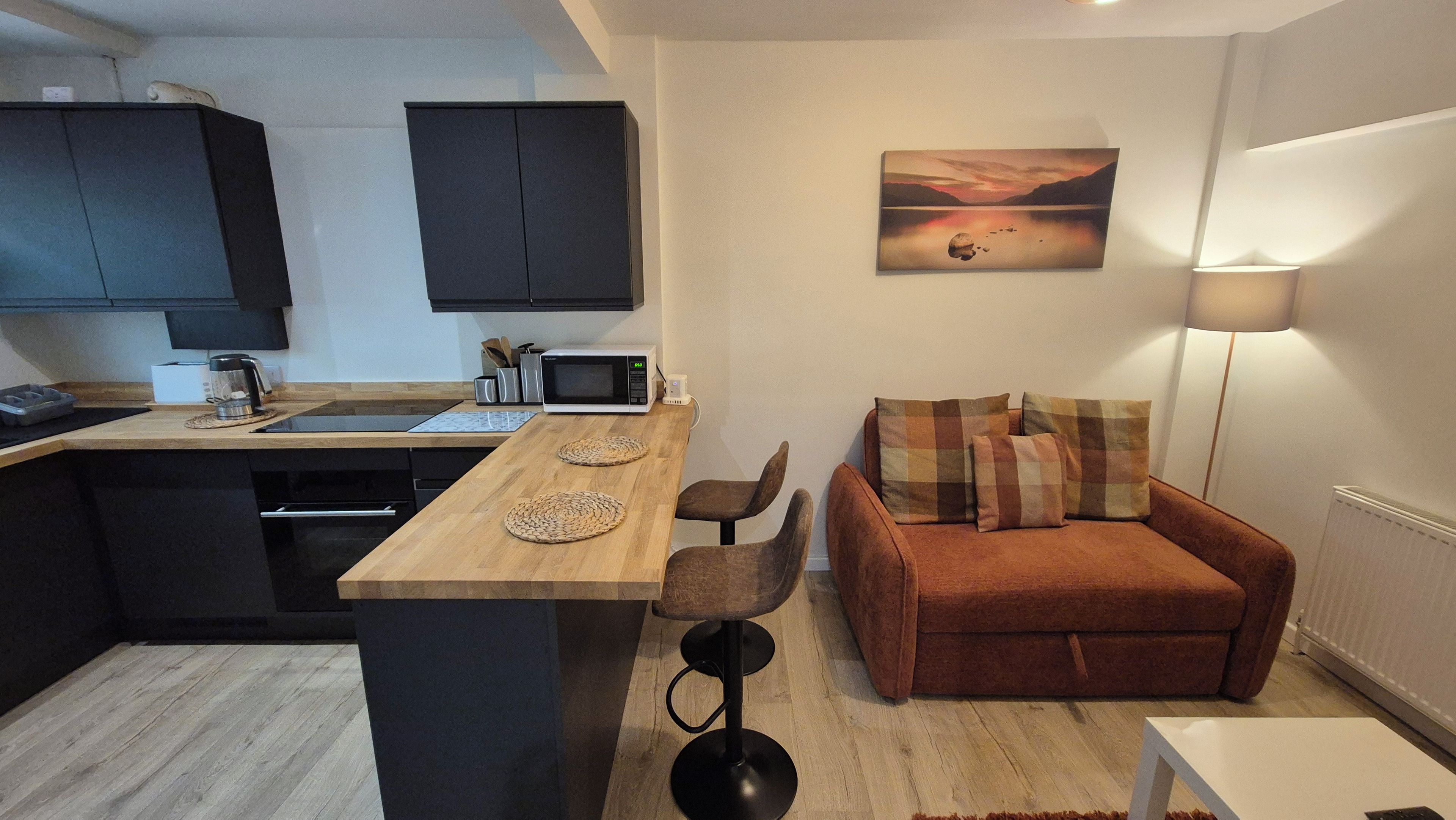 Un espace de vie compact comprenant un coin cuisine, un espace repas avec une table en bois, un canapé avec des coussins et un tableau accroché au mur.
