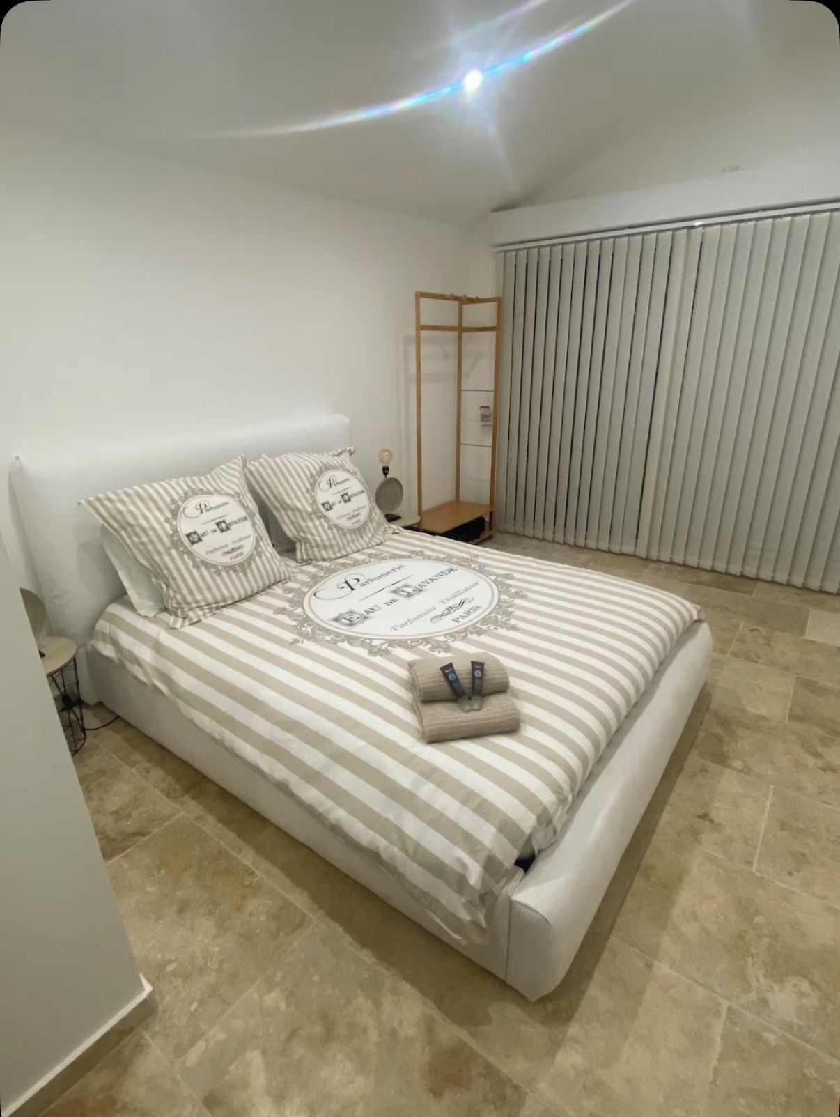 Een opgemaakt bed met gestreepte lakens en kussens, een nachtkastje met een lamp, en een raam met jaloezieën.
