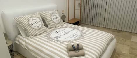 A neatly made bed with striped bedding and pillows, a bedside table with a lamp, and a window with blinds.