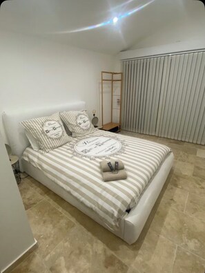 Een opgemaakt bed met gestreepte lakens en kussens, een nachtkastje met een lamp, en een raam met jaloezieën.