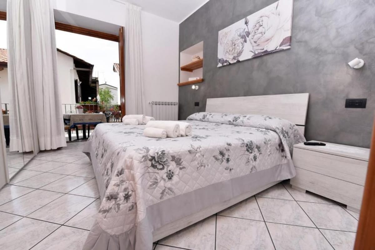 A bedroom with a large bed, a bedside table, a wall-mounted shelf, and a view of the outside.