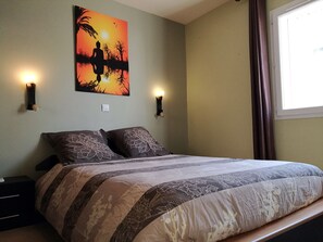 Uma cama com colcha estampada, duas luminárias fixadas na parede e uma pintura de pôr do sol na parede.