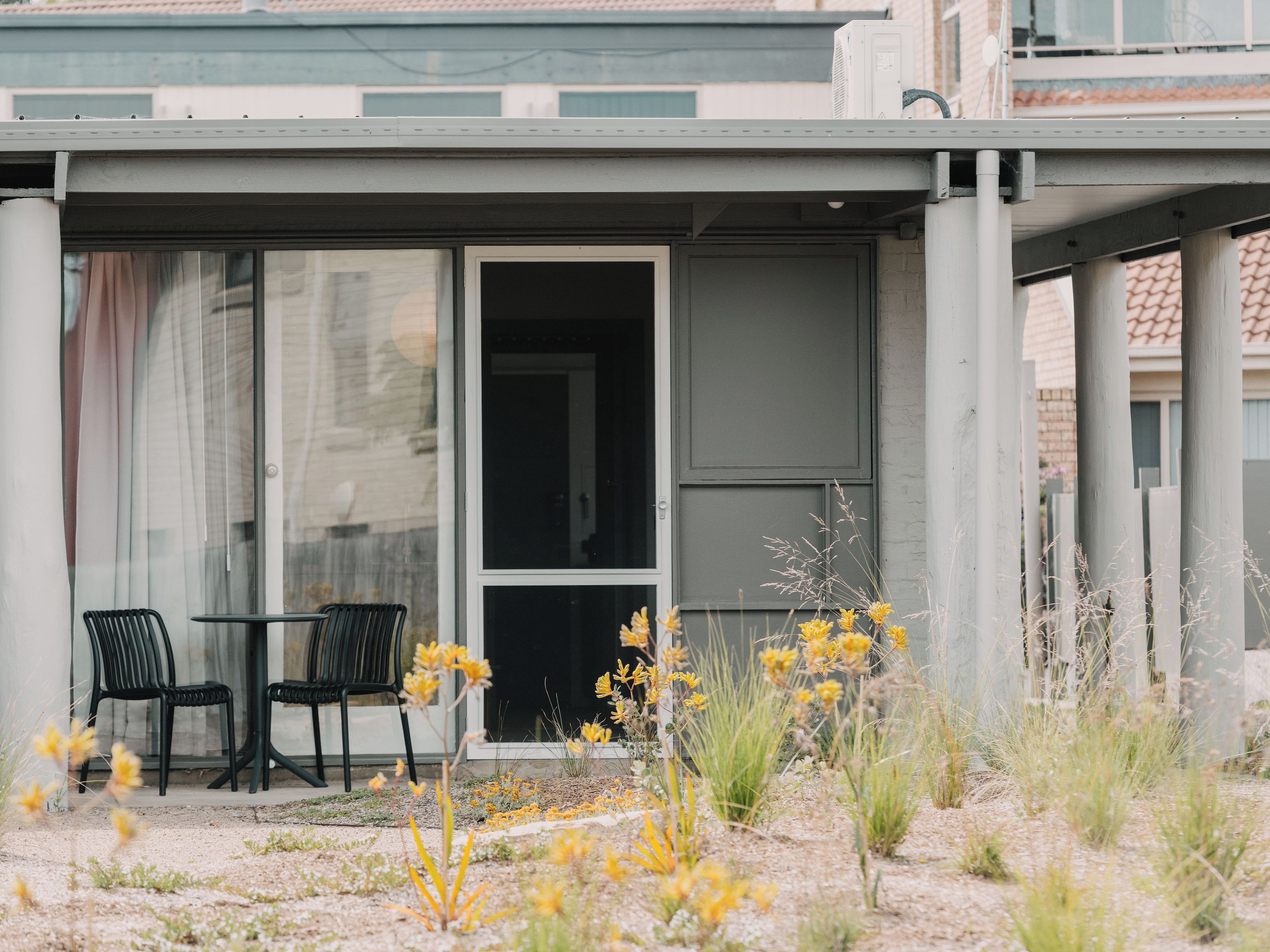 A covered outdoor seating area with two chairs and a table, surrounded by a garden with yellow flowers and grasses.