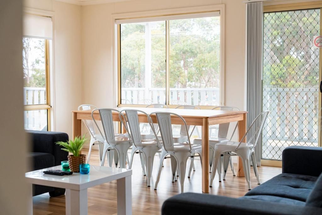 A dining area with a wooden table and white chairs, a small table with a plant and glasses, and a view through large windows.
