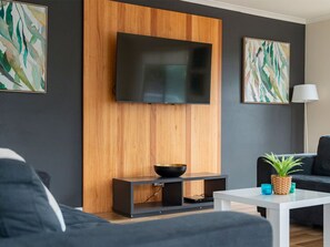 A modern living room with a flat-screen TV mounted on a wooden panel wall, a black sofa, a white coffee table, and a small potted plant. - The Black Dolphin Holiday Park (Merimbula)