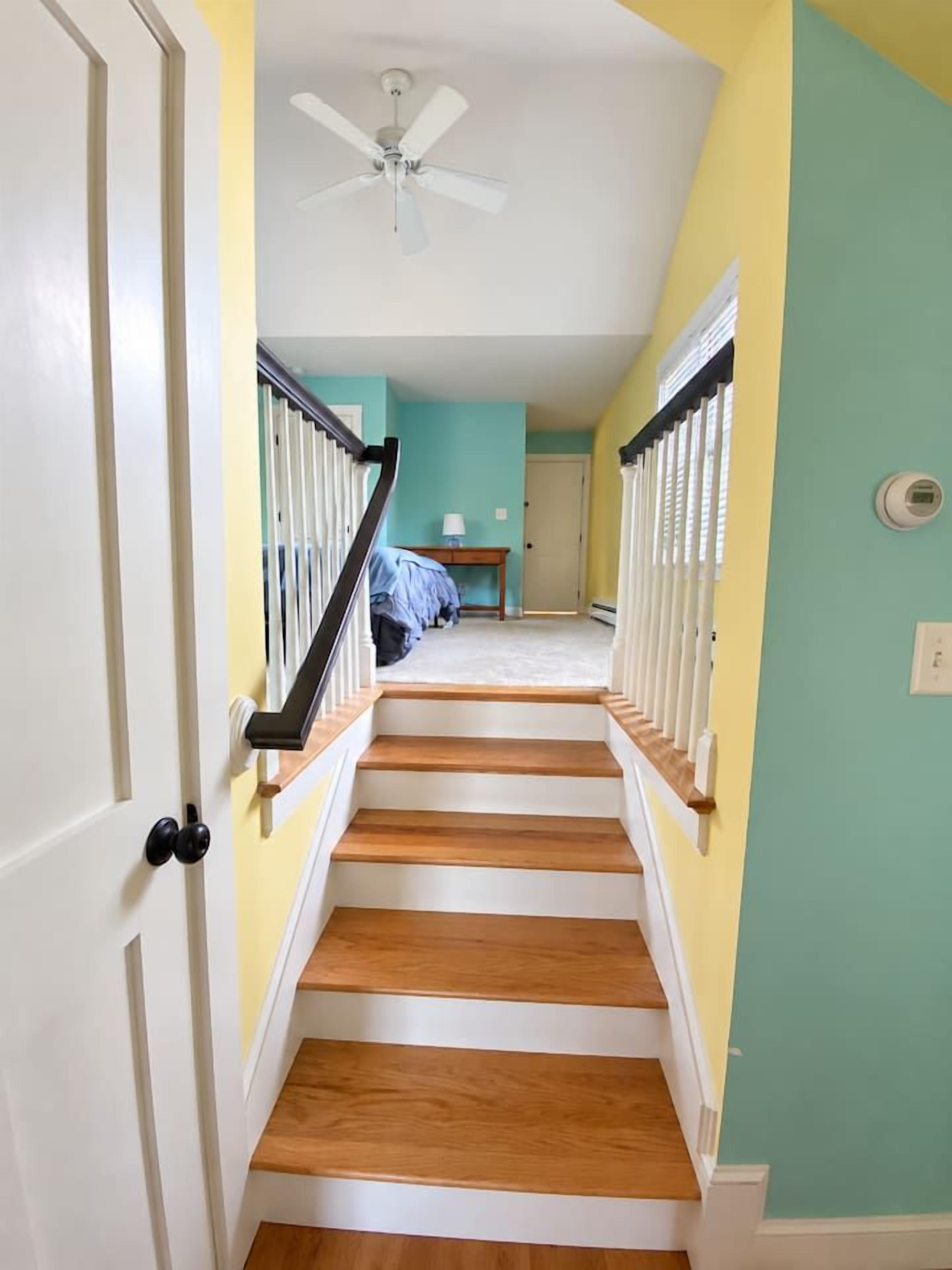 Un escalier avec des rampes blanches et des marches en bois menant à une pièce aux murs de couleur turquoise et dotée d’un ventilateur de plafond.