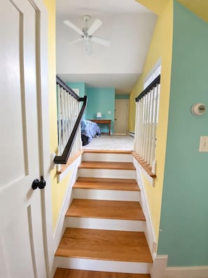 Un escalier avec des rampes blanches et des marches en bois menant à une pièce aux murs de couleur turquoise et dotée d’un ventilateur de plafond.