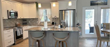 A modern kitchen with white cabinets, a stainless steel refrigerator, a large island with bar stools, and a dining area with a table and chairs.