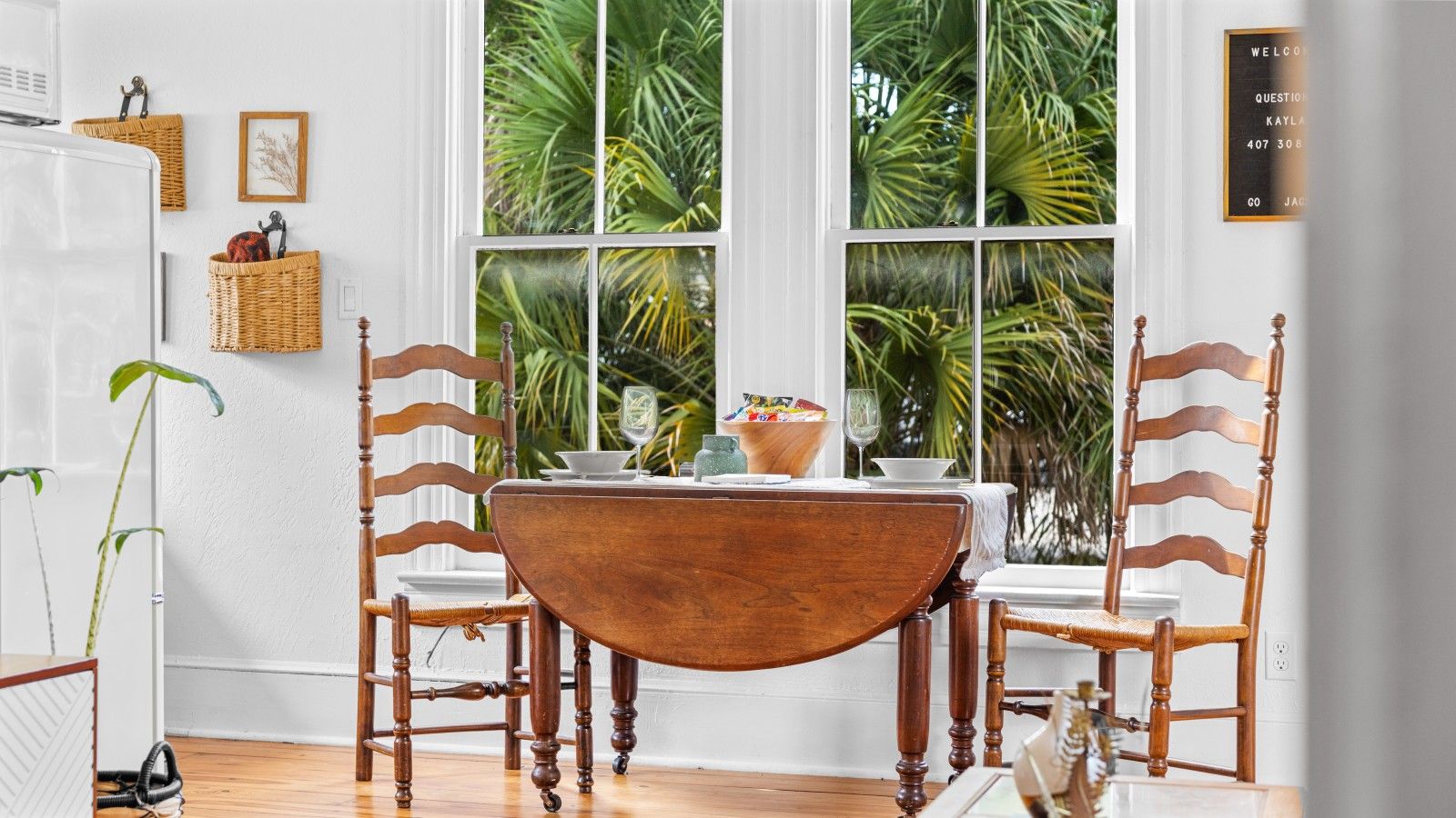 A dining area with a wooden table and chairs, a refrigerator, and a window with a view of palm trees.