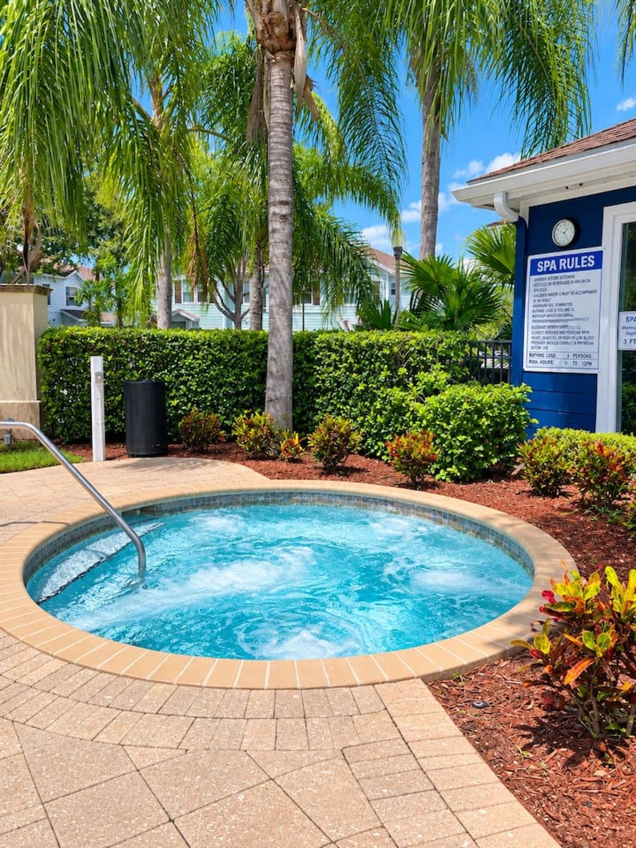 A hot tub surrounded by a landscaped garden with palm trees and a hedge, and a sign indicating SPA RULES.