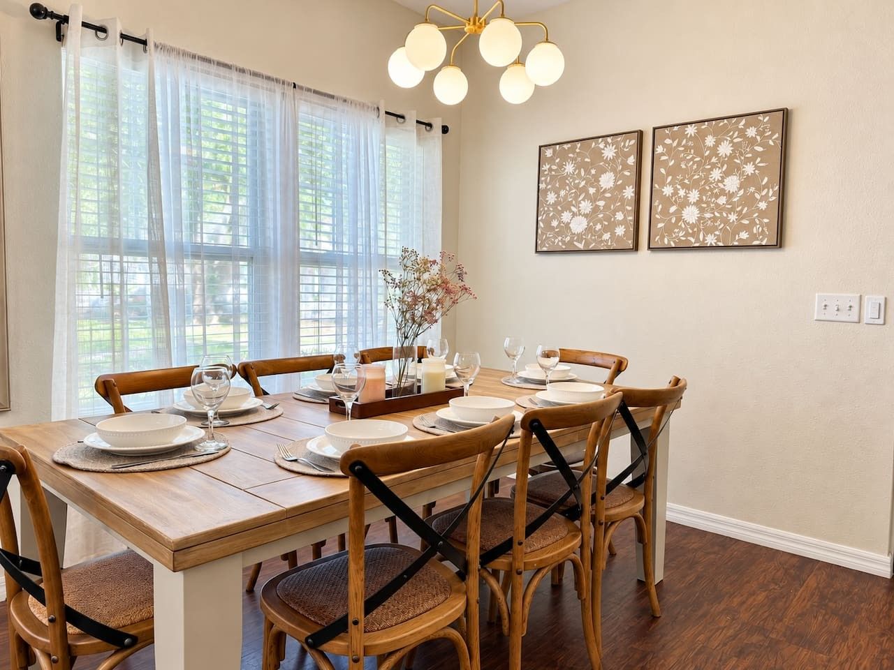 A dining area with a wooden table set for four, surrounded by chairs, with a window and two framed floral artworks on the wall.