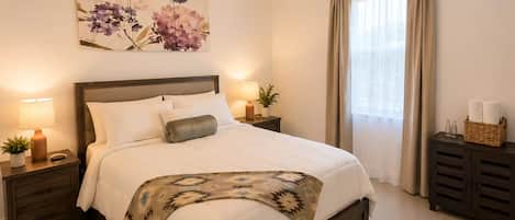 A bedroom with a bed, bedside tables, a nightstand, a floor lamp, a wall-mounted light fixture, a window with curtains, and a decorative wall piece.