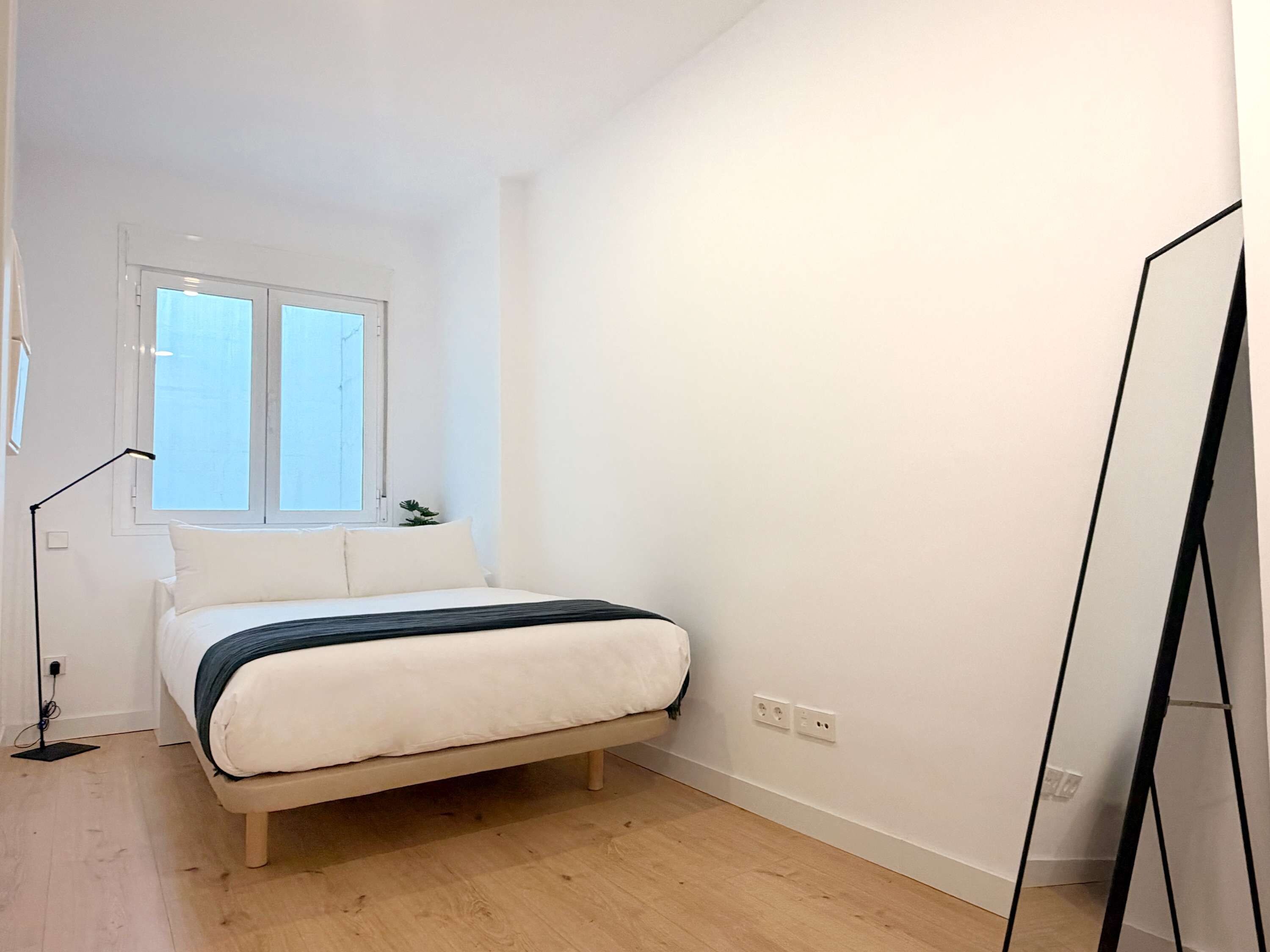 Ein Schlafzimmer mit einem Bett, einem Fenster, einer Stehlampe und einem schwarzen Gegenstand rechts.