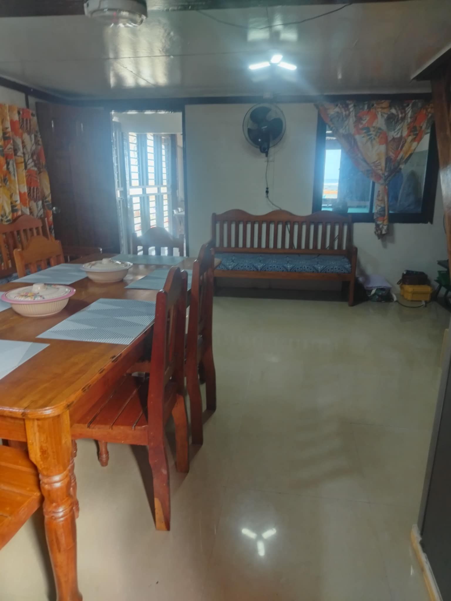A dining area with a wooden table, chairs, and a bench. There is a fan on the wall and a window with curtains.