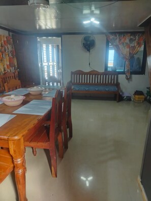 A dining area with a wooden table, chairs, and a bench. There is a fan on the wall and a window with curtains. - GUMOBAO STAYCATION HOUSE (Baganga)