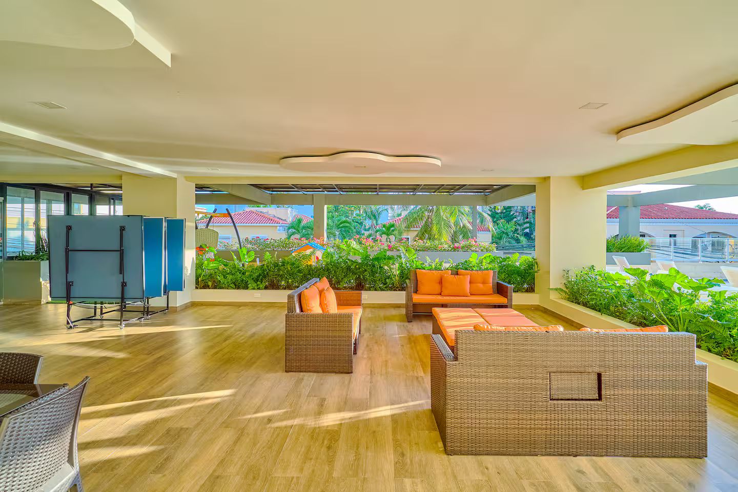 A spacious indoor area with a wooden floor, a seating area with wicker furniture, and a view of a garden with plants and a building in the background.