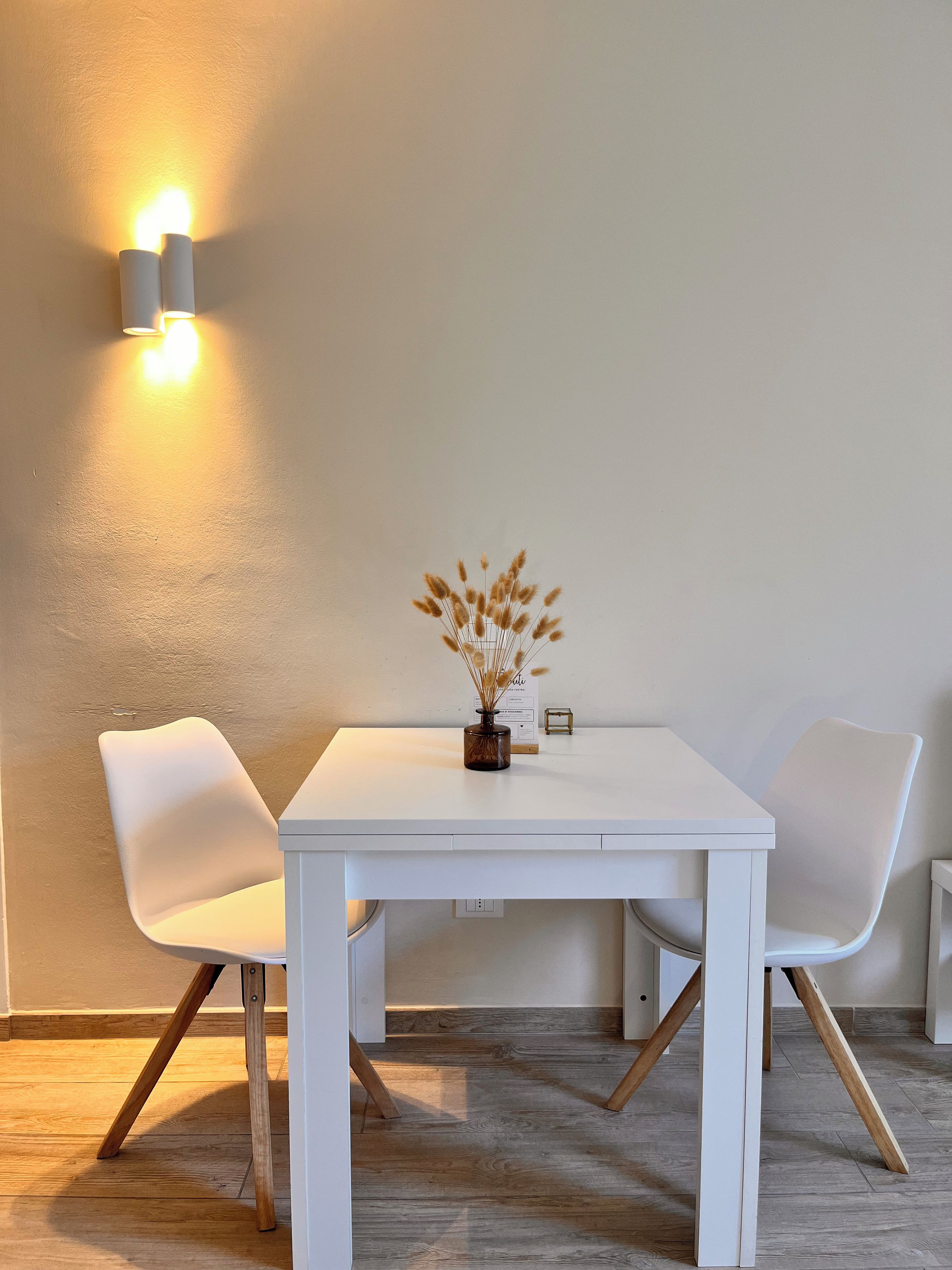 A white table with two chairs, a vase with dried plants, and a wall-mounted light fixture.