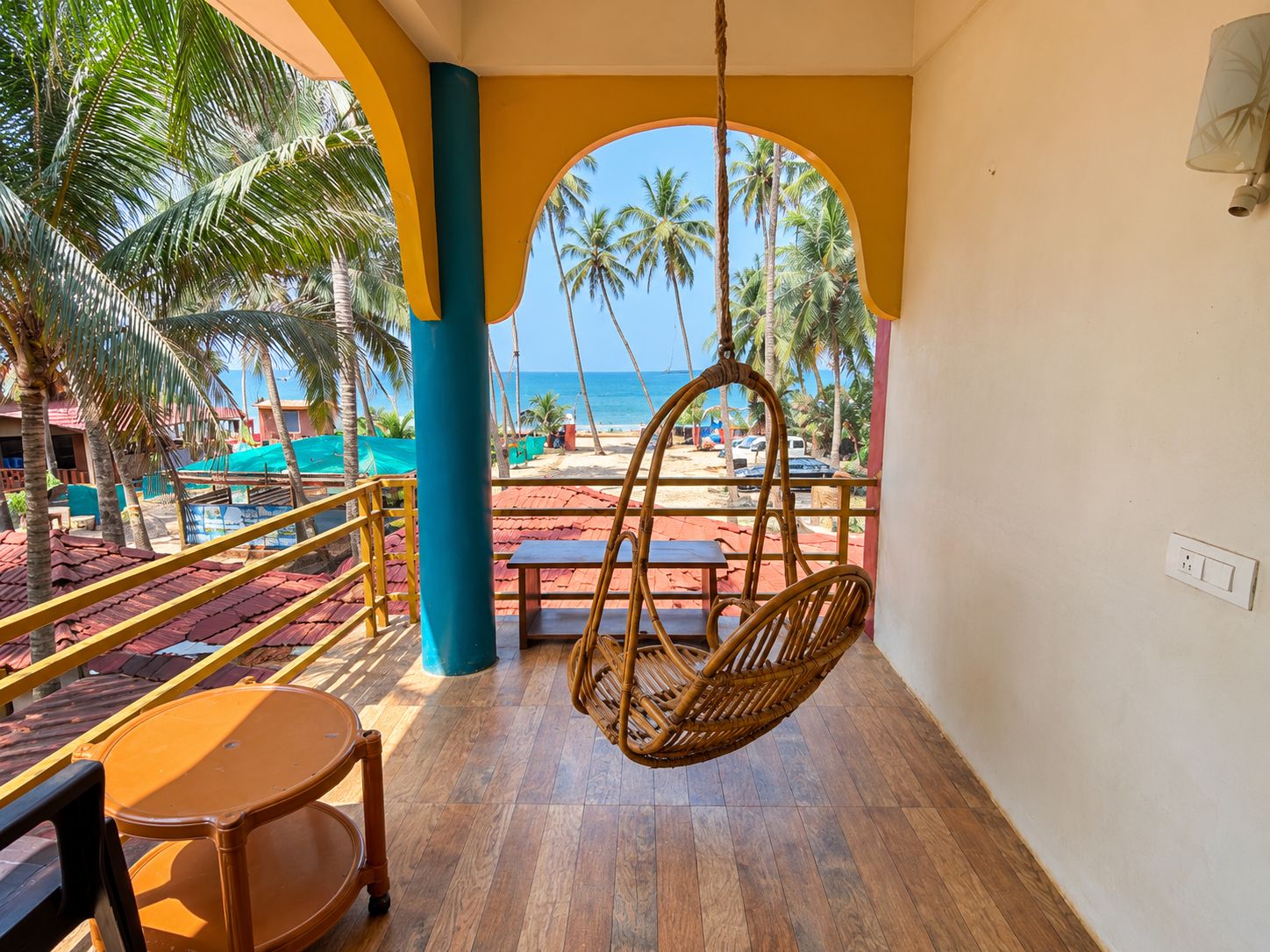 A balcony with a hanging chair, wooden flooring, and a view of the beach and palm trees.