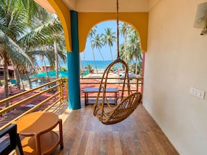 Un balcón con una hamaca colgante, suelo de madera y vistas a la playa y palmeras.