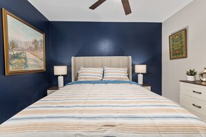Une chambre à coucher comprenant un lit, deux tables de chevet, une commode, un ventilateur de plafond et un tableau accroché au mur.