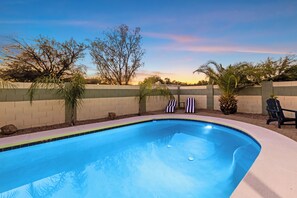 Une piscine dont le bord est recouvert de carreaux, entourée d’un petit muret en briques, avec quelques arbres et un coucher de soleil en arrière-plan.