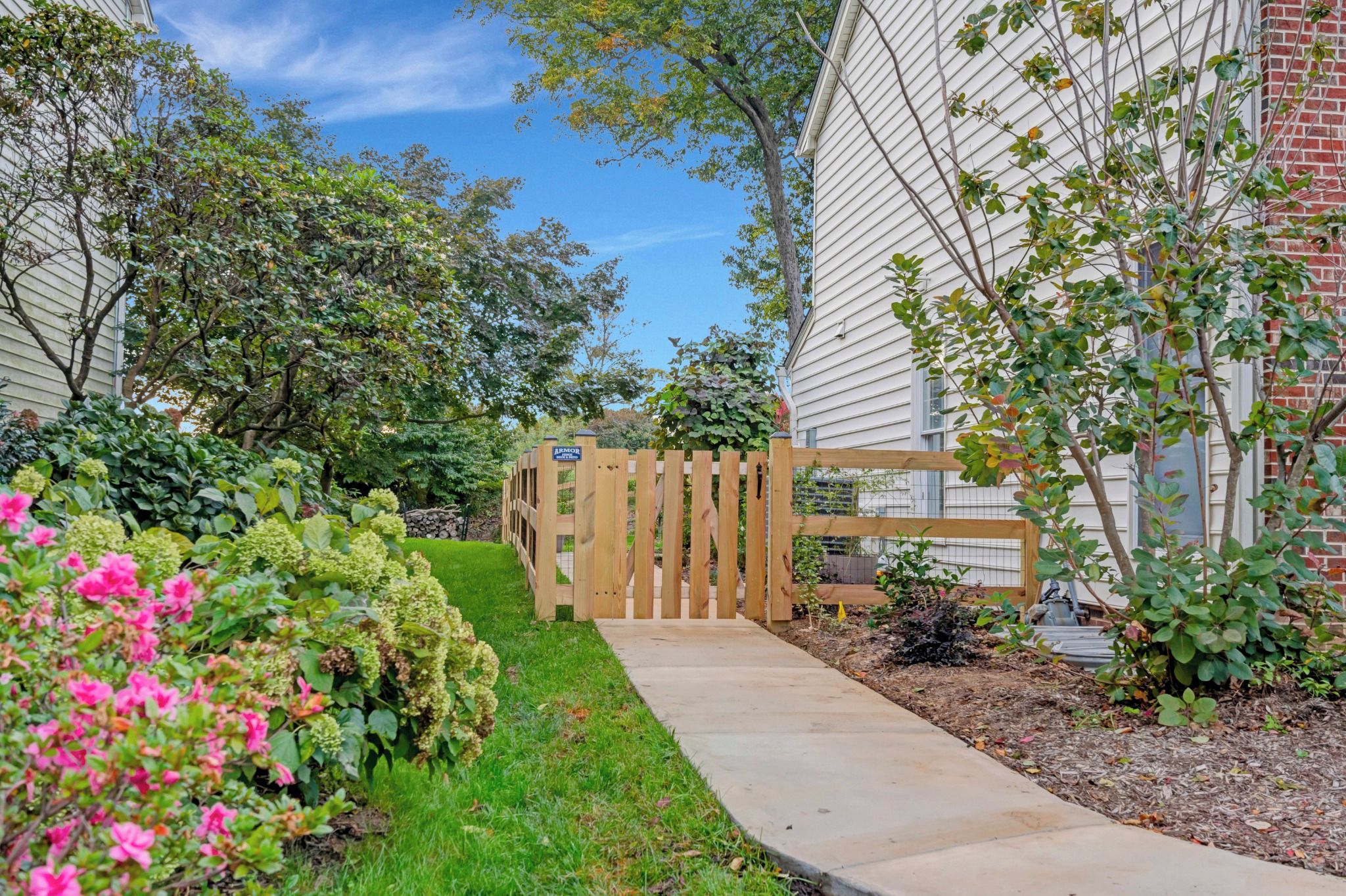 A pathway leading to a wooden gate with a sign, surrounded by lush greenery and flowering plants.