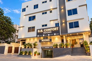 Ein mehrstöckiges Hotel namens 'B.B. Comfort Rooms' mit einem Schild, das Annehmlichkeiten wie Klimaanlage, kostenloses WLAN, Warmwasser und Parkplätze anzeigt.