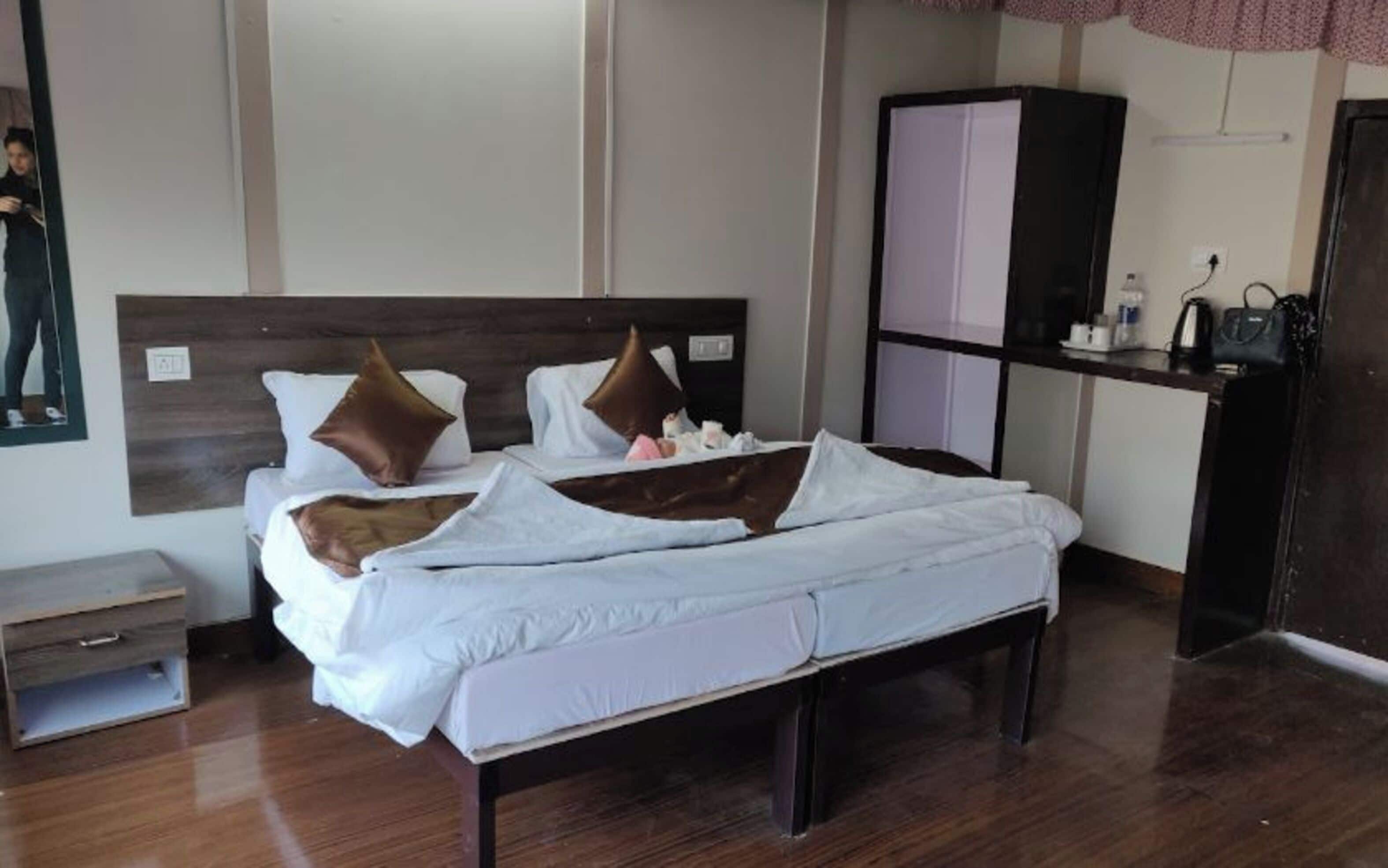 A neatly made bed with white bedding and brown pillows, a wooden nightstand with a drawer, a dark wooden wardrobe, and a small table with toiletries.