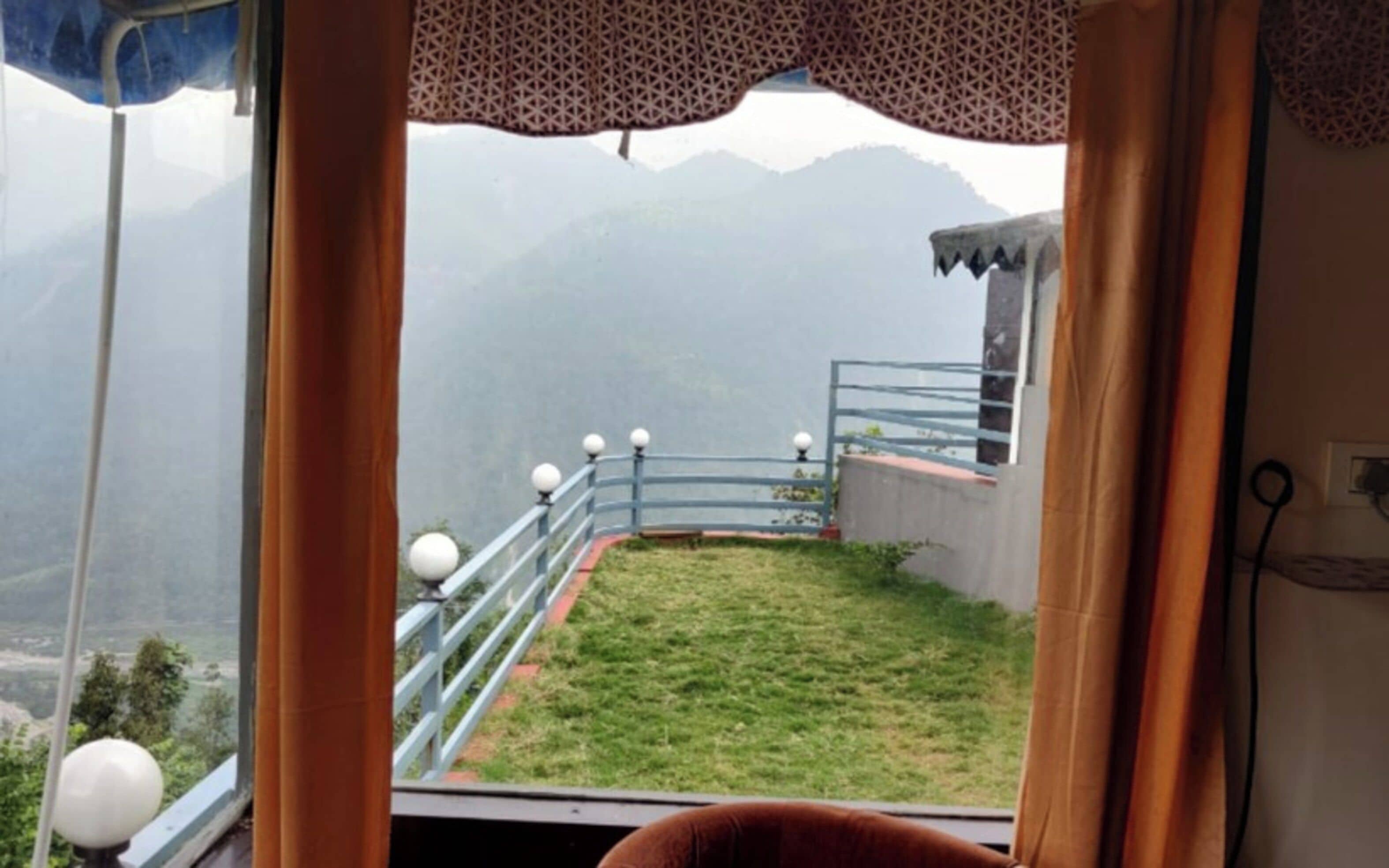 Vista desde una ventana con balcón, sobre un área verde y montañas distantes.