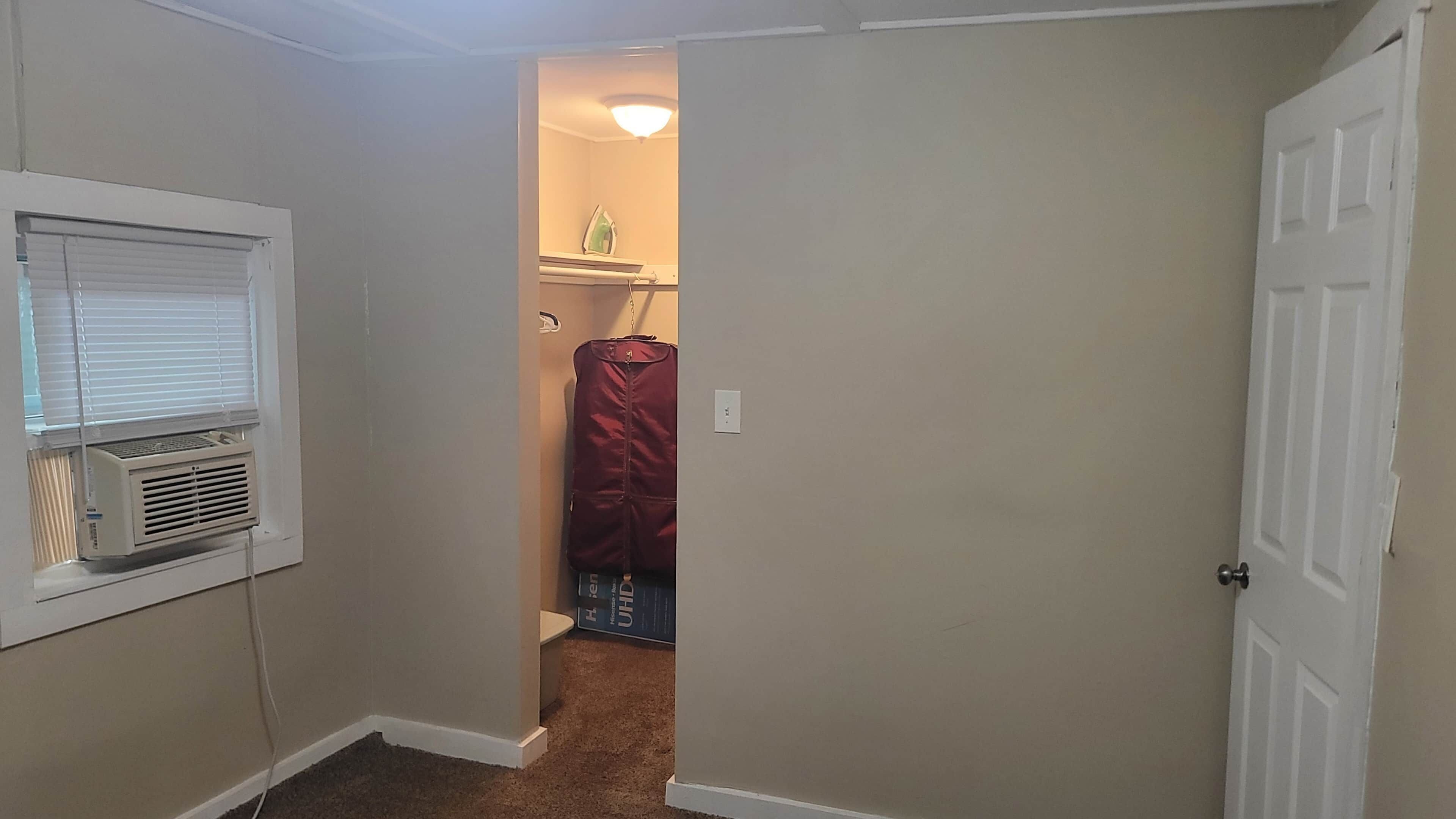 Ein schmaler Flur mit einer weißen Tür rechts, einer weißen Wand mit einem Fenster links und einer roten Jacke, die an der Wand hängt.