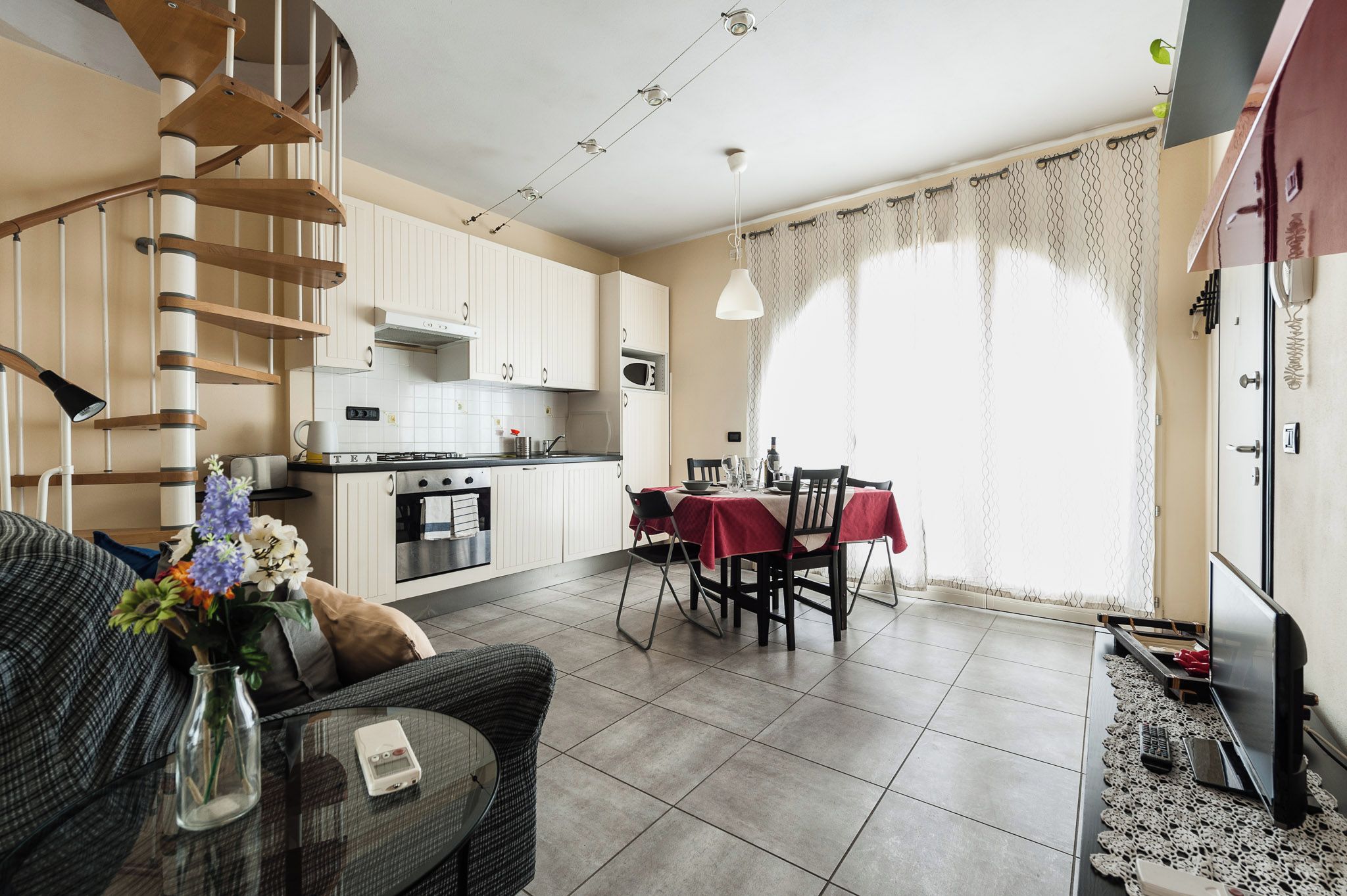 A modern kitchen with white cabinets, a dining area with a red tablecloth, and a staircase with wooden railings.